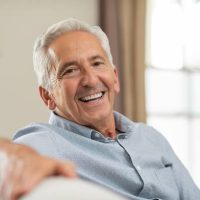 Portrait of happy senior man smiling at home. Old man relaxing on sofa and looking at camera. Portrait of elderly man enjoying retirement.